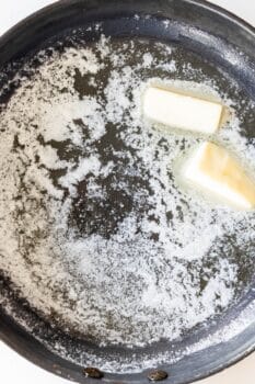 Two pieces of butter melting in a black skillet on a stove, perfect for starting delicious Butter Poached Potatoes.