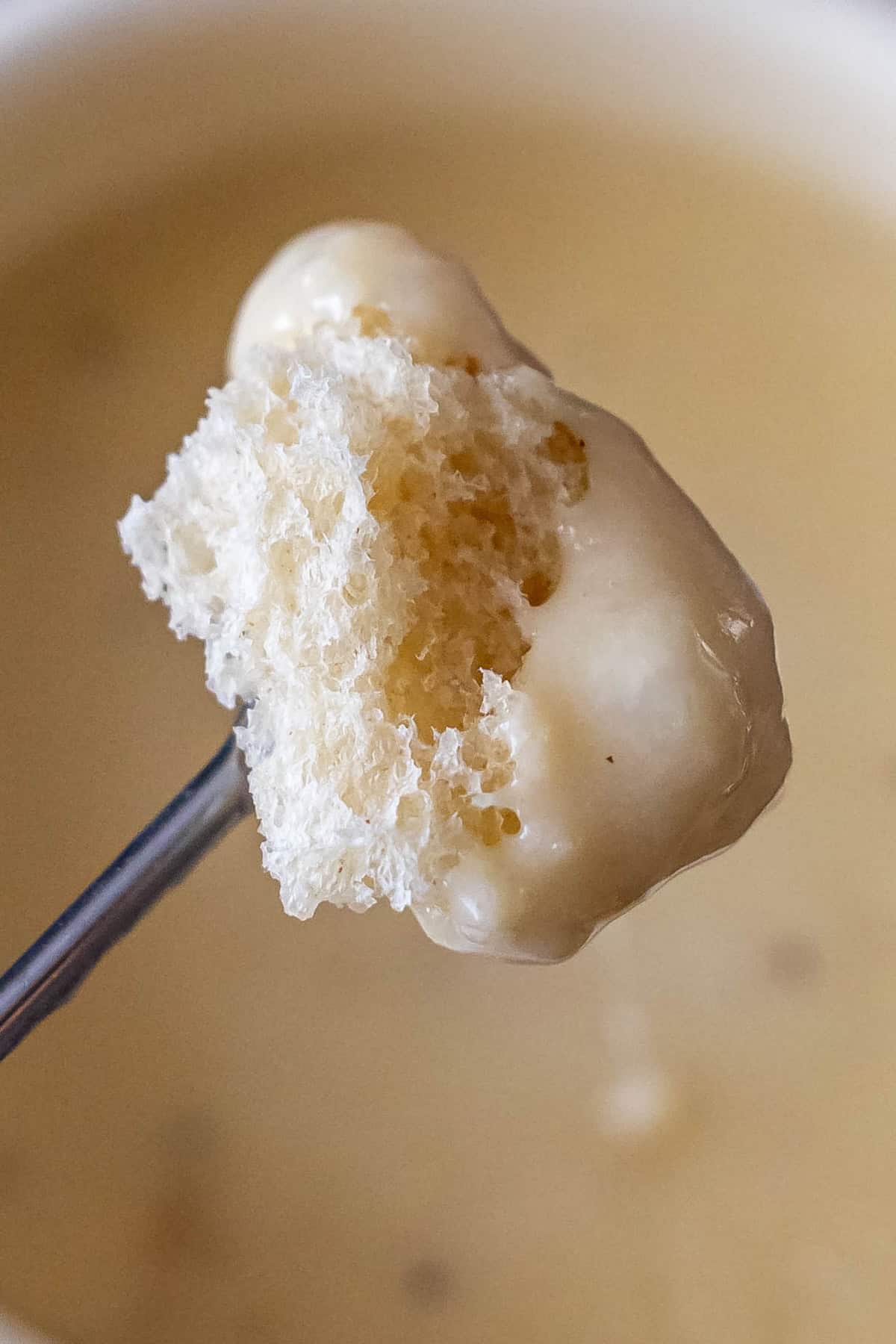 A close-up of a fork holding a piece of bread dipped in creamy cheese fondue, with the bowl of the best cheese fondue blurred in the background.