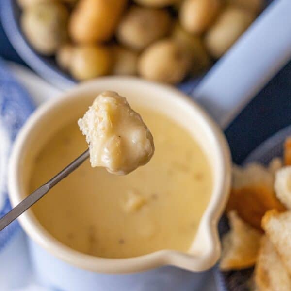 A piece of bread dipped in the best cheese fondue is held above a pot of fondue, with extra bread pieces and potatoes visible in the background.