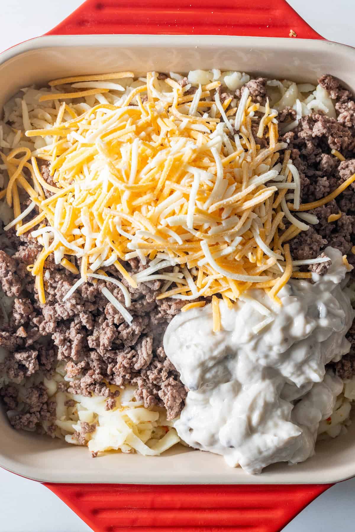 A baking dish with layers of cooked ground beef, shredded cheese, and a creamy white sauce—this cheesy casserole is ready to be mixed or baked to golden perfection.