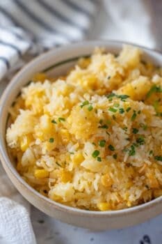 A bowl of Corn Rice, mixed with corn and garnished with chopped herbs, sits on a table with a striped cloth in the background.