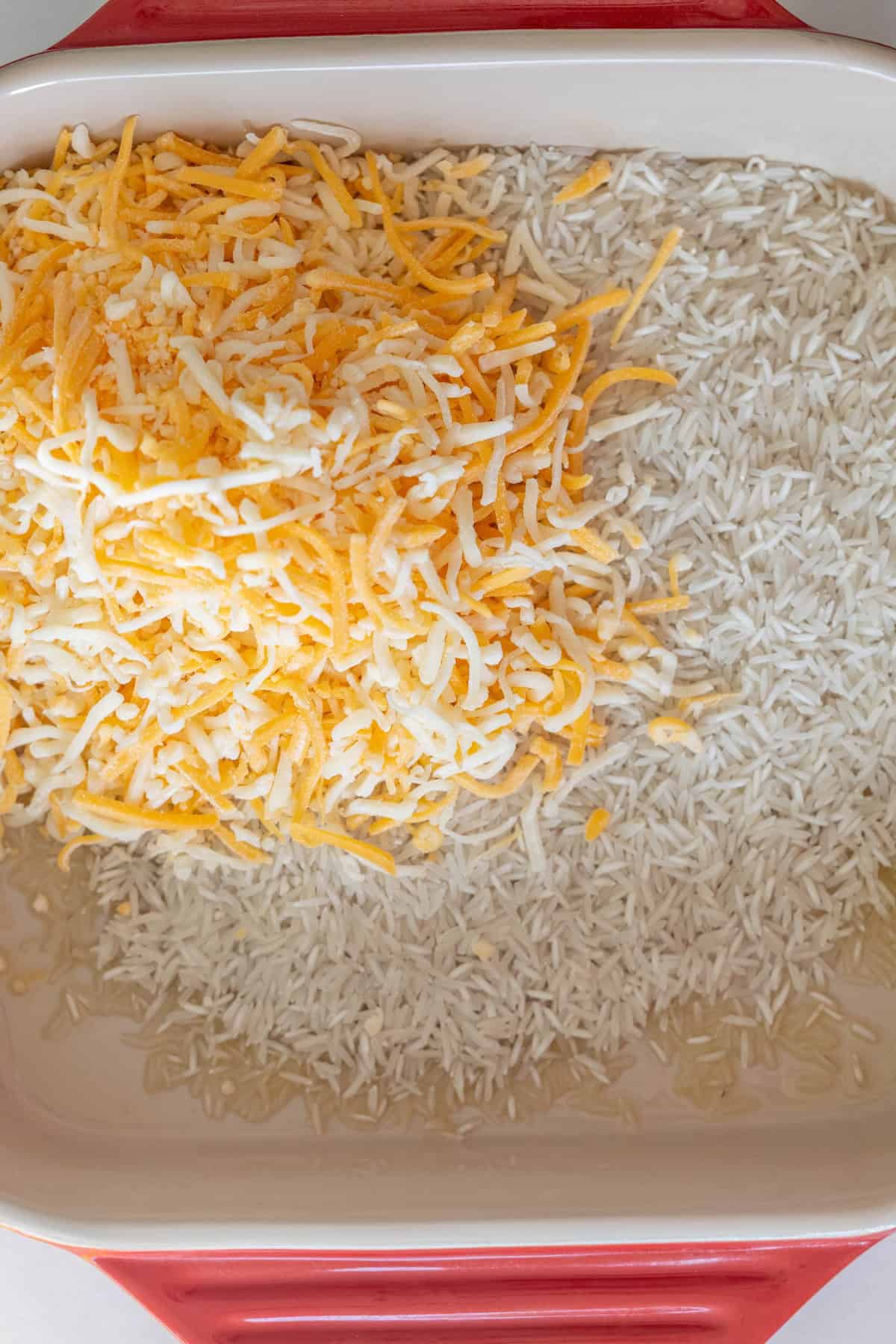 Uncooked white rice and a pile of shredded yellow and white cheese in a red baking dish, ready to be transformed into a delicious baked casserole.