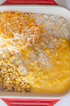 A casserole dish containing shredded cheese, shredded hash browns, creamed corn, whole corn, and breadcrumbs is shown before mixing—perfect for a baked cheesy corn rice casserole.