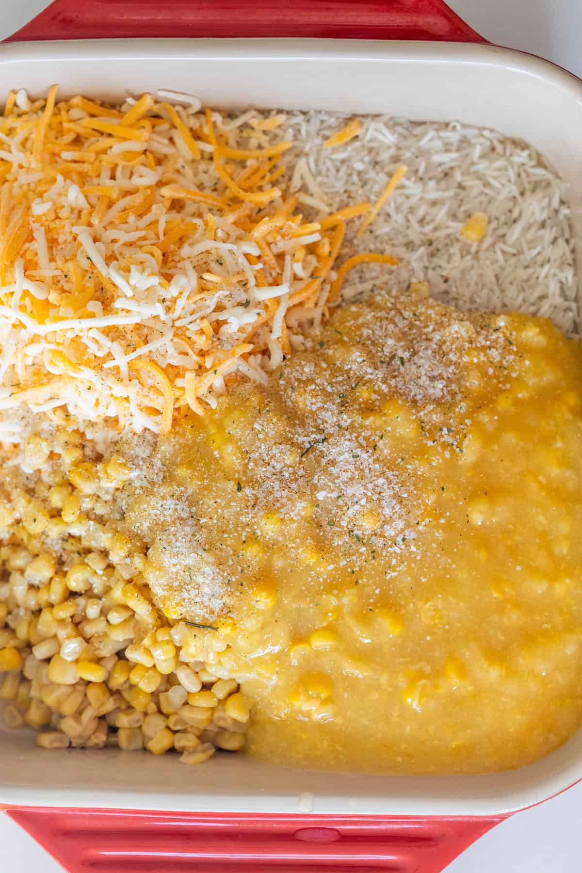 A casserole dish containing shredded cheese, shredded hash browns, creamed corn, whole corn, and breadcrumbs is shown before mixing&mdash;perfect for a baked cheesy corn rice casserole.