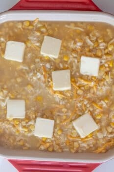 A casserole dish containing a mixture of rice, corn, shredded cheese, and water, topped with butter slices—this Cheesy Corn Rice Casserole is ready to be baked.