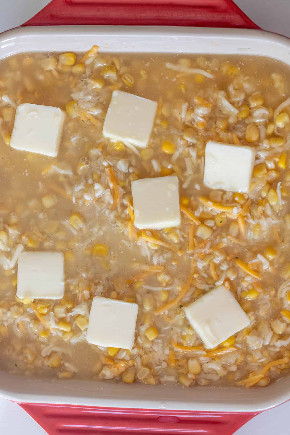 A casserole dish containing a mixture of rice, corn, shredded cheese, and water, topped with butter slices&mdash;this Cheesy Corn Rice Casserole is ready to be baked.