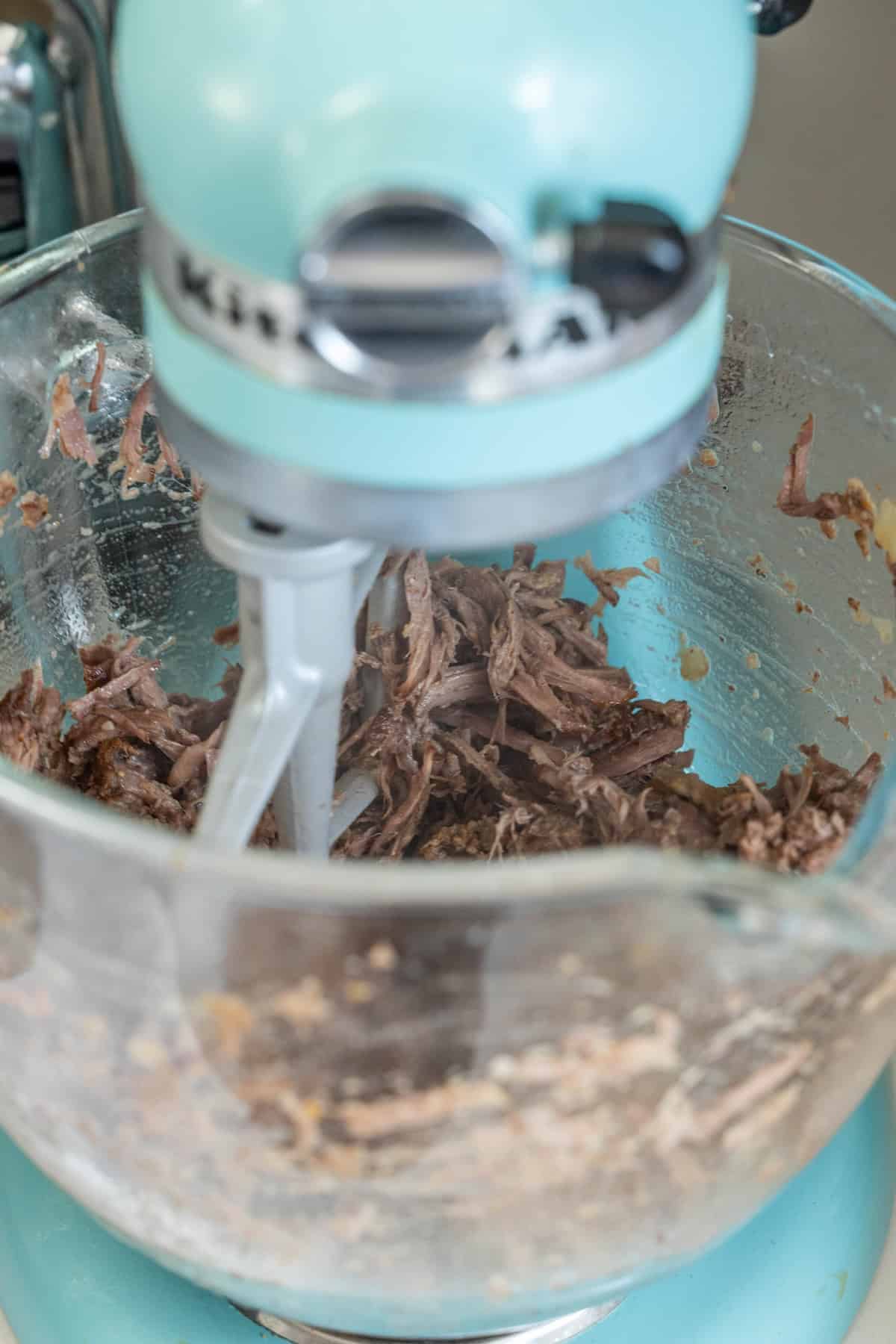 A stand mixer with a paddle attachment is mixing shredded Piri Piri meat in a glass bowl.