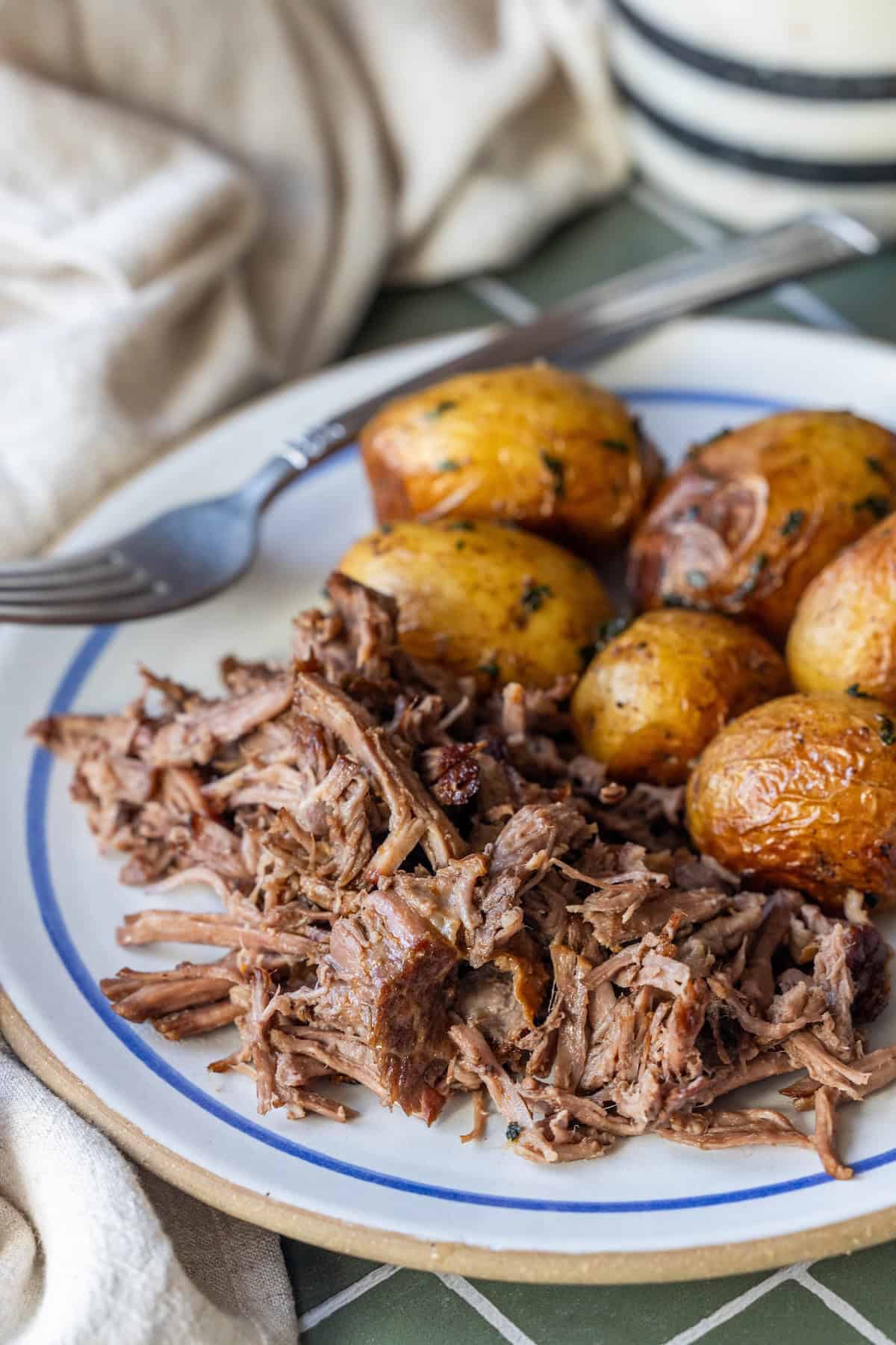 Plate of tender Slow Cooker Chuck Roast and roasted baby potatoes, served with a fork on a green tiled surface and a beige napkin nearby.