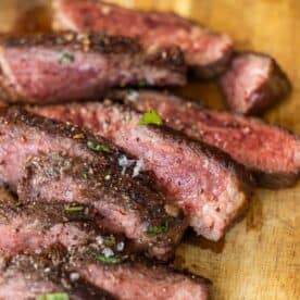 Several slices of medium-rare Denver Steak topped with coarse salt and herbs are arranged on a wooden cutting board.