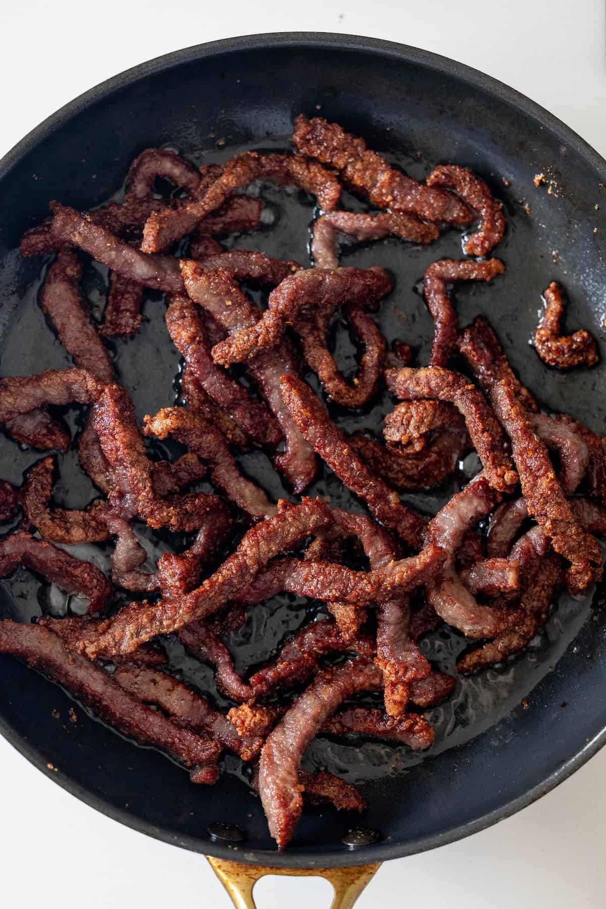 A black skillet containing cooked, browned beef strips with a crispy texture on a white surface&mdash;perfect for an easy, takeout style Mongolian Beef at home.
