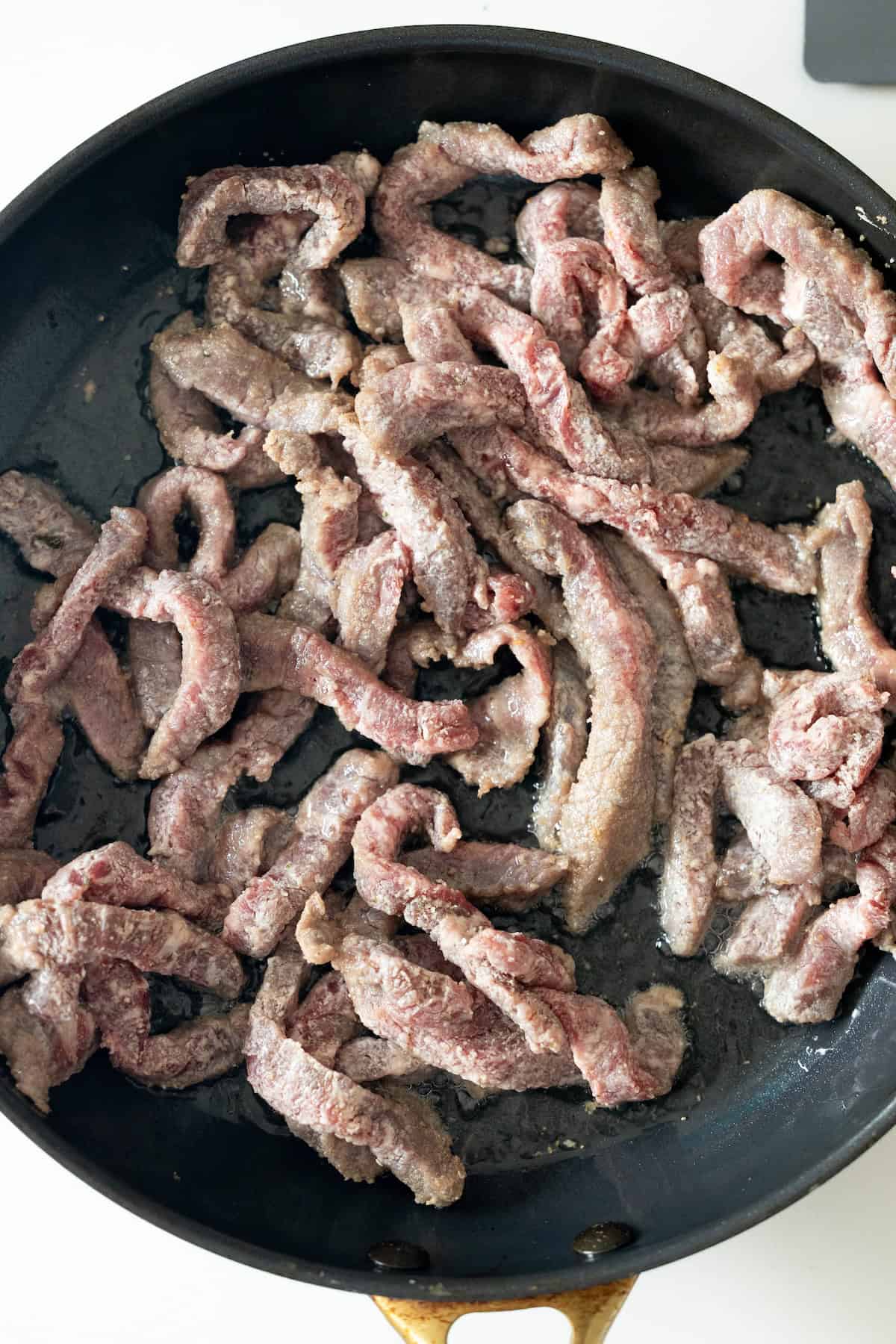 Strips of raw beef cooking in a black skillet on a stovetop, perfect for making easy Mongolian Beef that tastes just like your favorite takeout.