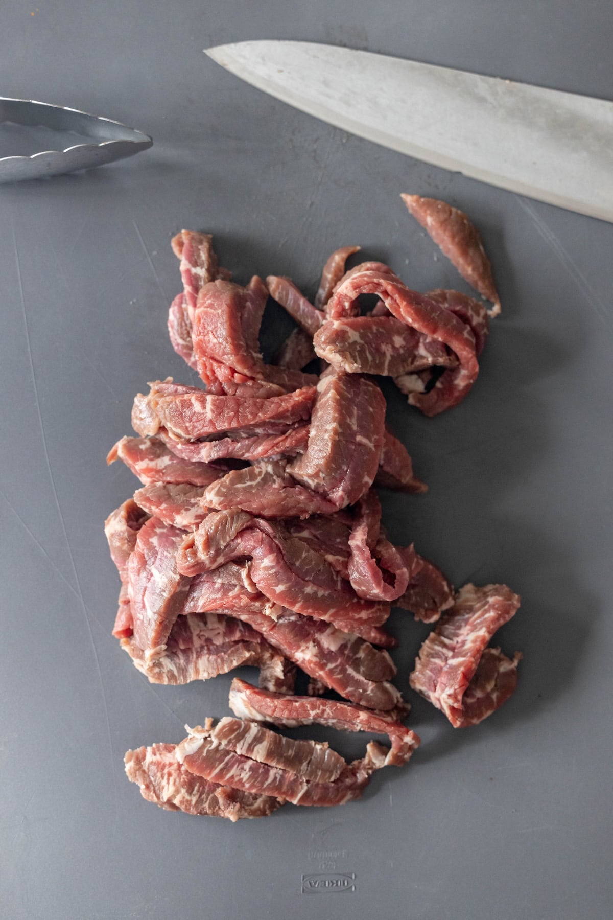 Sliced raw beef strips on a gray cutting board, ready for easy Mongolian Beef&mdash;just like your favorite takeout style! A large knife and metal tongs are nearby for quick meal prep.