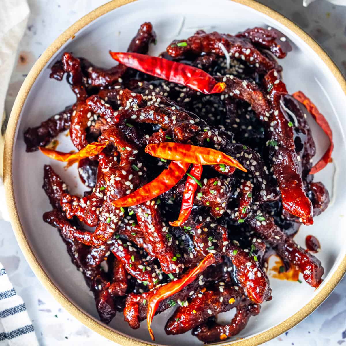 A bowl of easy, takeout-style Mongolian Beef featuring crispy fried beef strips coated in a dark sauce, garnished with red dried chili peppers and sprinkled with sesame seeds.