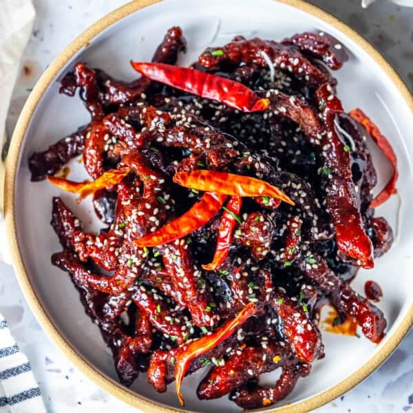 A bowl of easy, takeout-style Mongolian Beef featuring crispy fried beef strips coated in a dark sauce, garnished with red dried chili peppers and sprinkled with sesame seeds.