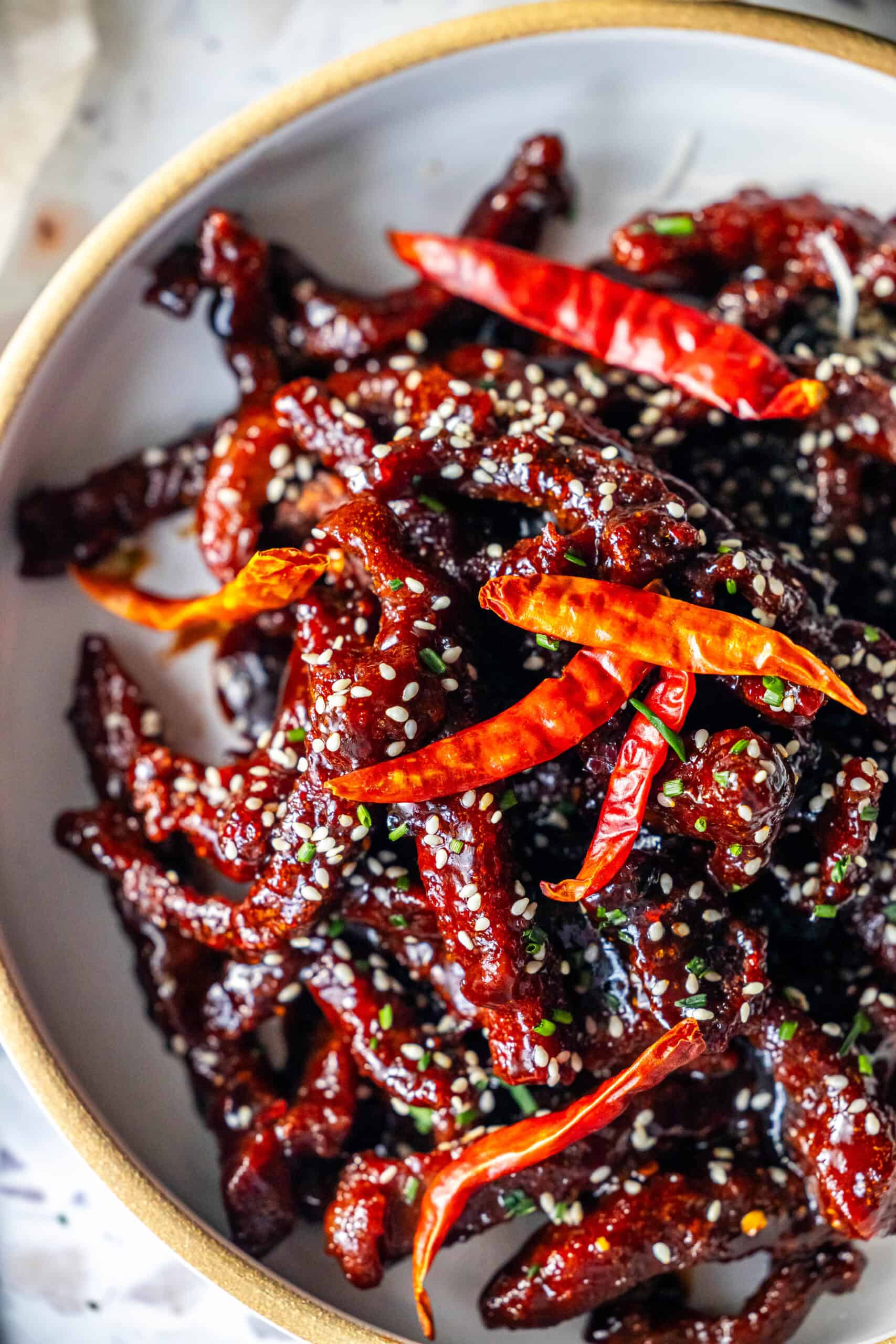 A plate of easy, takeout style Mongolian Beef features crispy beef strips in a dark sauce, garnished with sesame seeds, dried red chilies, and chopped green onions.