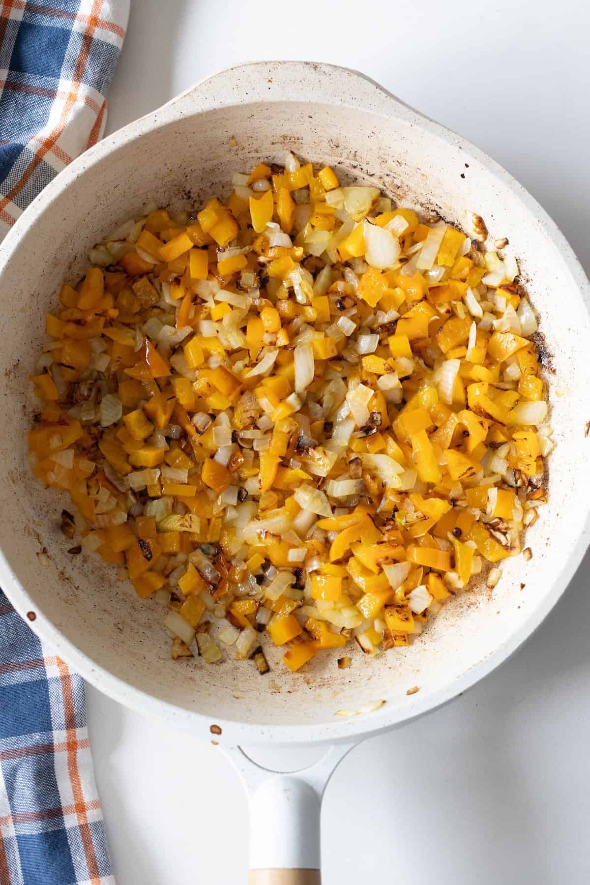 Chopped yellow bell peppers and onions are saut&eacute;ing in a white skillet, perfect for starting an Old Fashioned Ham and Beans recipe, with a blue and orange plaid cloth nearby on a white surface.