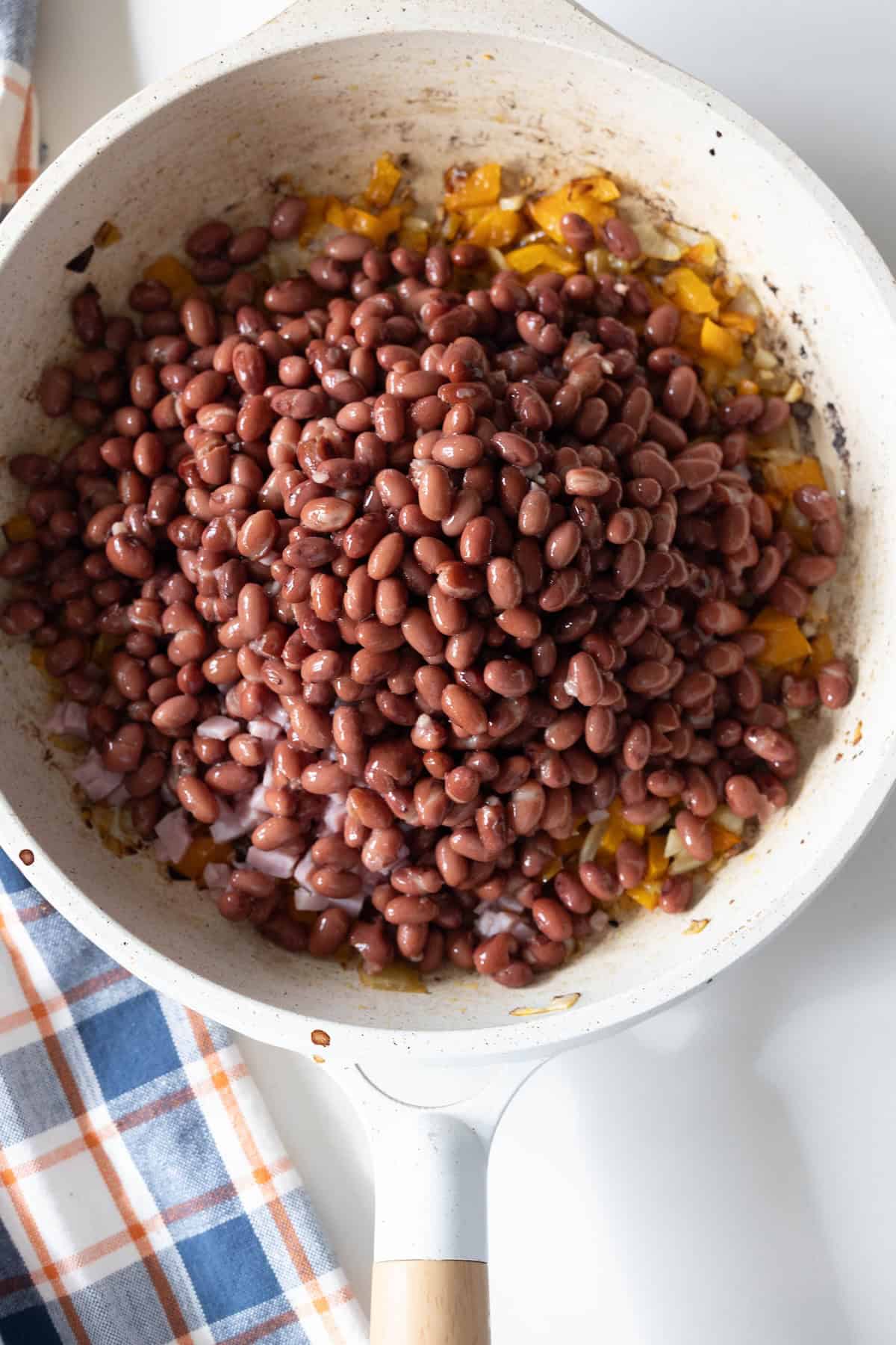 A white skillet filled with cooked black beans, chopped yellow bell pepper, and onions sits on a white surface next to a plaid cloth, evoking the comforting flavors of an Old Fashioned Ham and Beans recipe.