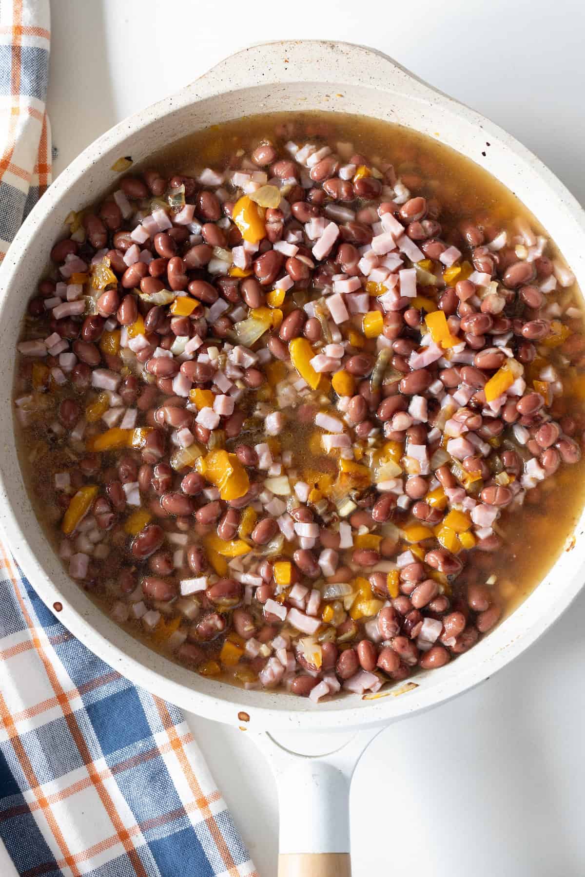 A white skillet containing Old Fashioned Ham and Beans with diced ham, cooked beans, chopped onions, and yellow bell peppers in a broth sits next to a plaid cloth.