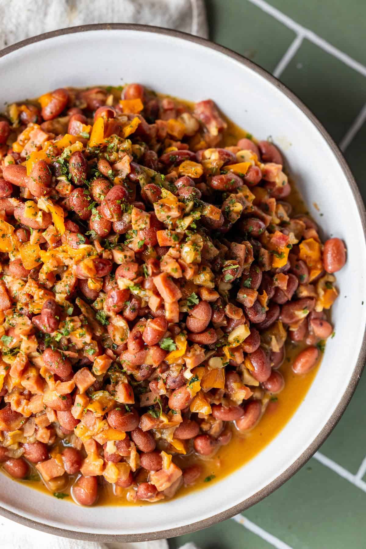 A bowl of old-fashioned pinto beans mixed with chopped vegetables and herbs in a light broth sits on a green tiled surface.