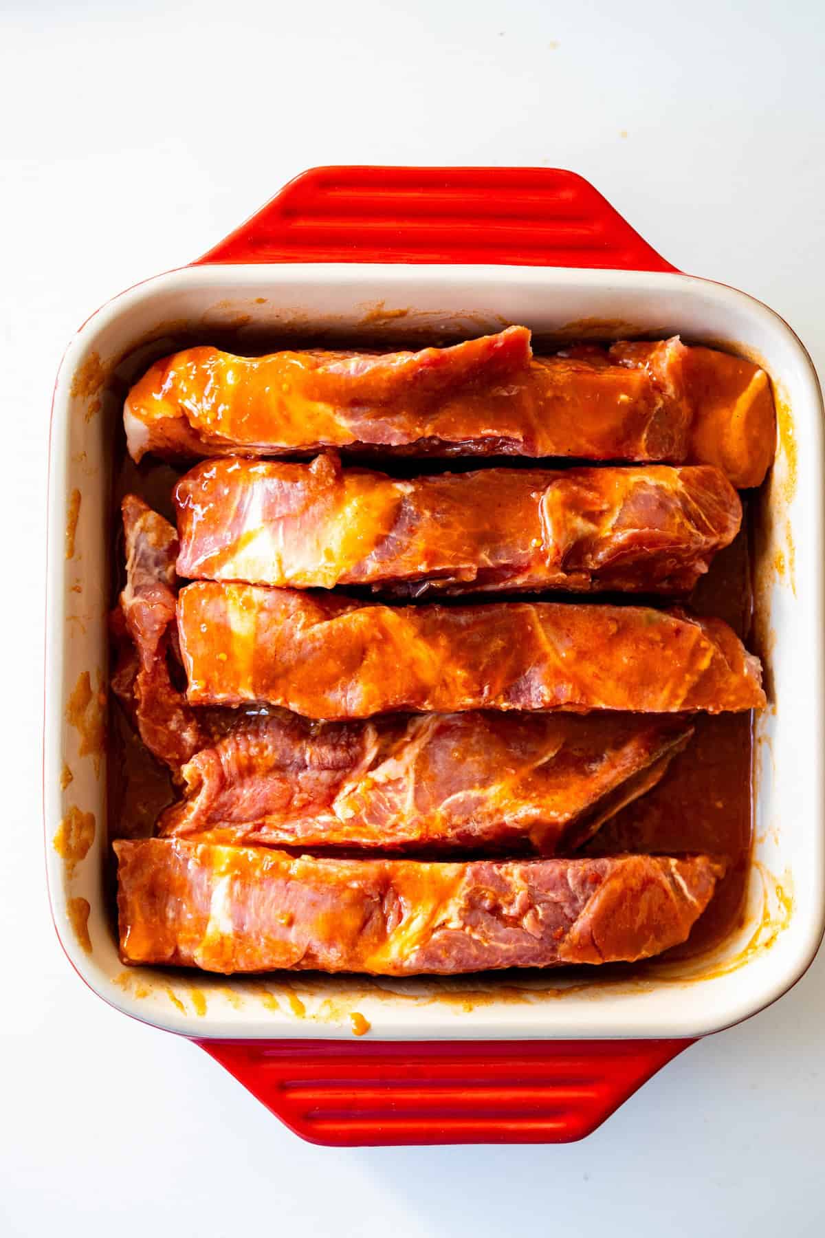 Raw country style ribs coated in marinade are arranged in a red and white baking dish on a white surface, ready to become flavorful roasted ribs.