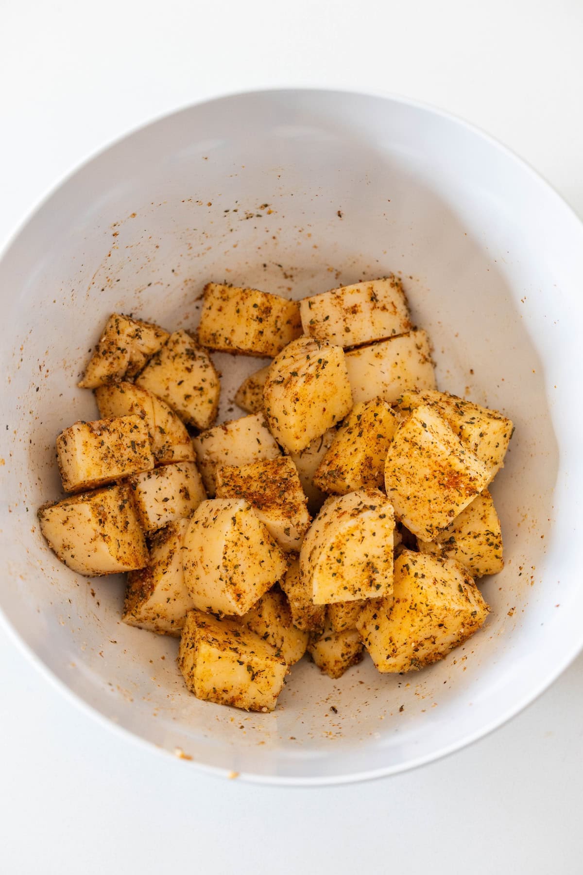 Cubed potatoes coated with spices in a white mixing bowl, perfect alongside Baked Polenta Bites for a flavorful snack or side.