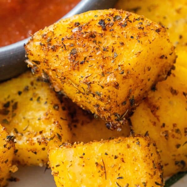 Close-up of several seasoned, crispy yellow Baked Polenta Bites with visible herbs and spices, served next to a bowl of red dipping sauce.