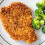 A golden-brown chicken cutlet, inspired by a classic Schnitzel recipe, is served alongside steamed broccoli on a white plate with a green rim.