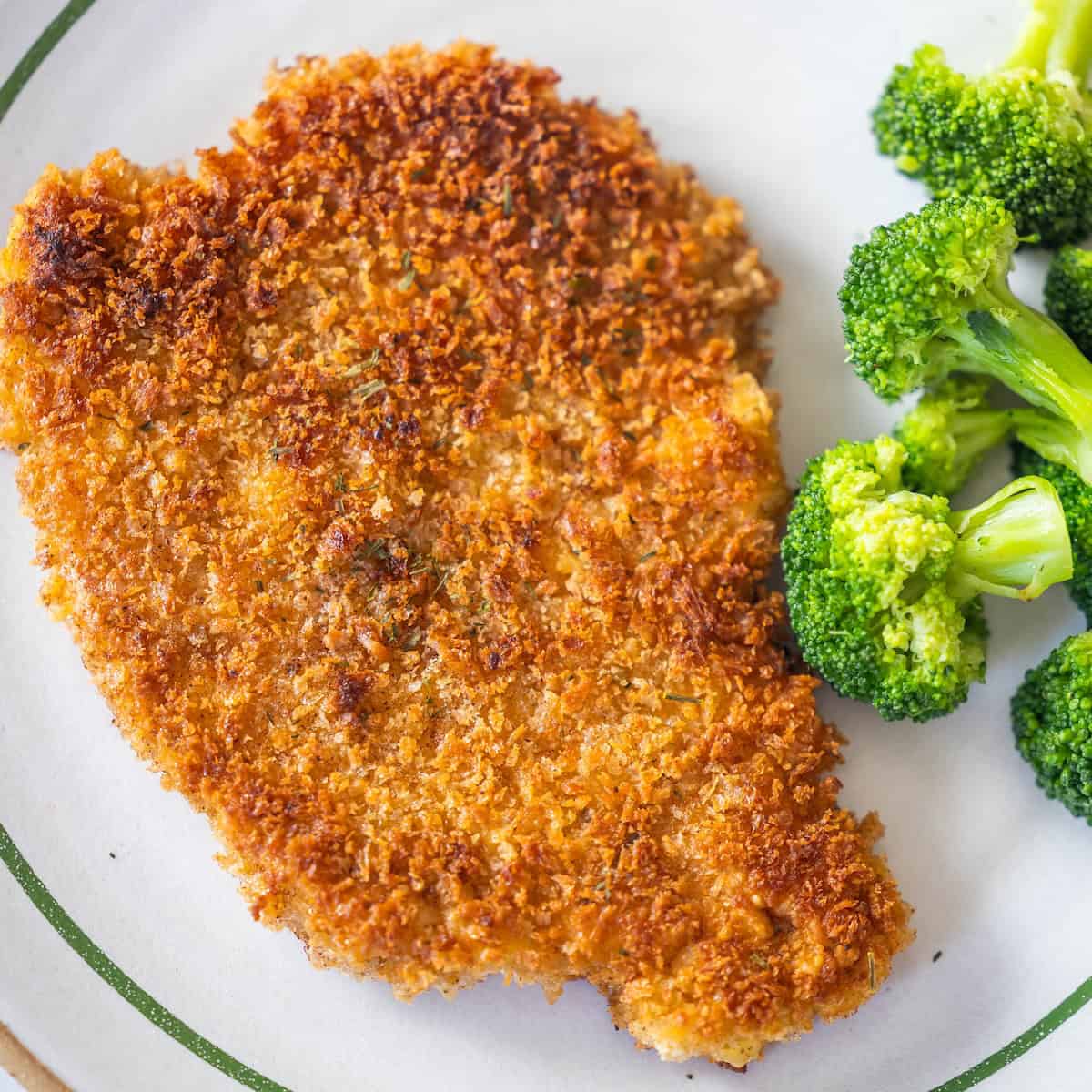 A golden-brown chicken cutlet, inspired by a classic Schnitzel recipe, is served alongside steamed broccoli on a white plate with a green rim.