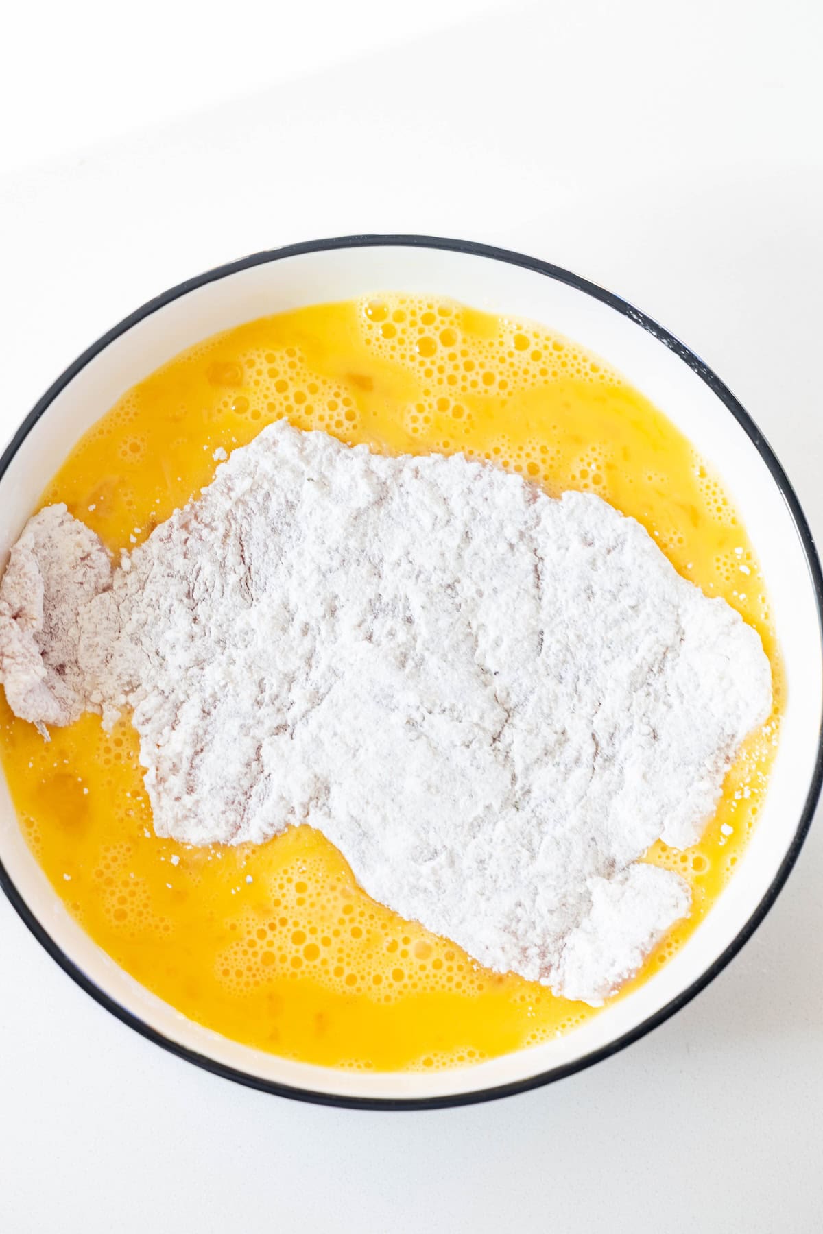 A bowl with beaten eggs and a piece of flour-coated meat being dipped into the mixture, perfect for preparing a classic Pork Schnitzel recipe, on a white surface.