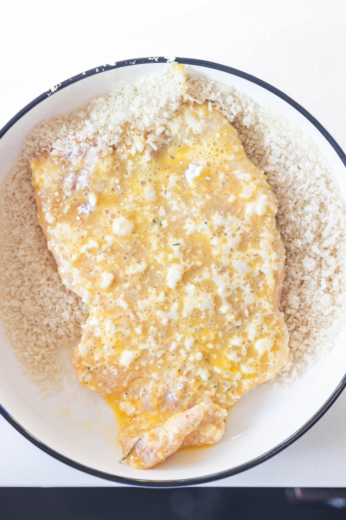 A raw breaded chicken cutlet coated in beaten egg is placed on a bed of panko breadcrumbs in a white shallow bowl, similar to the method used in a classic Schnitzel recipe.