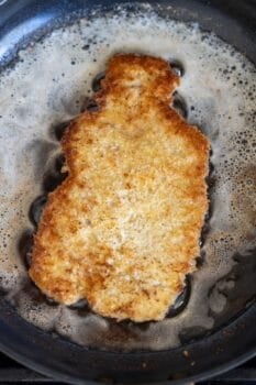 A breaded cutlet is being fried in a pan with bubbling oil and butter, revealing the golden brown crust typical of a classic Pork Schnitzel recipe.