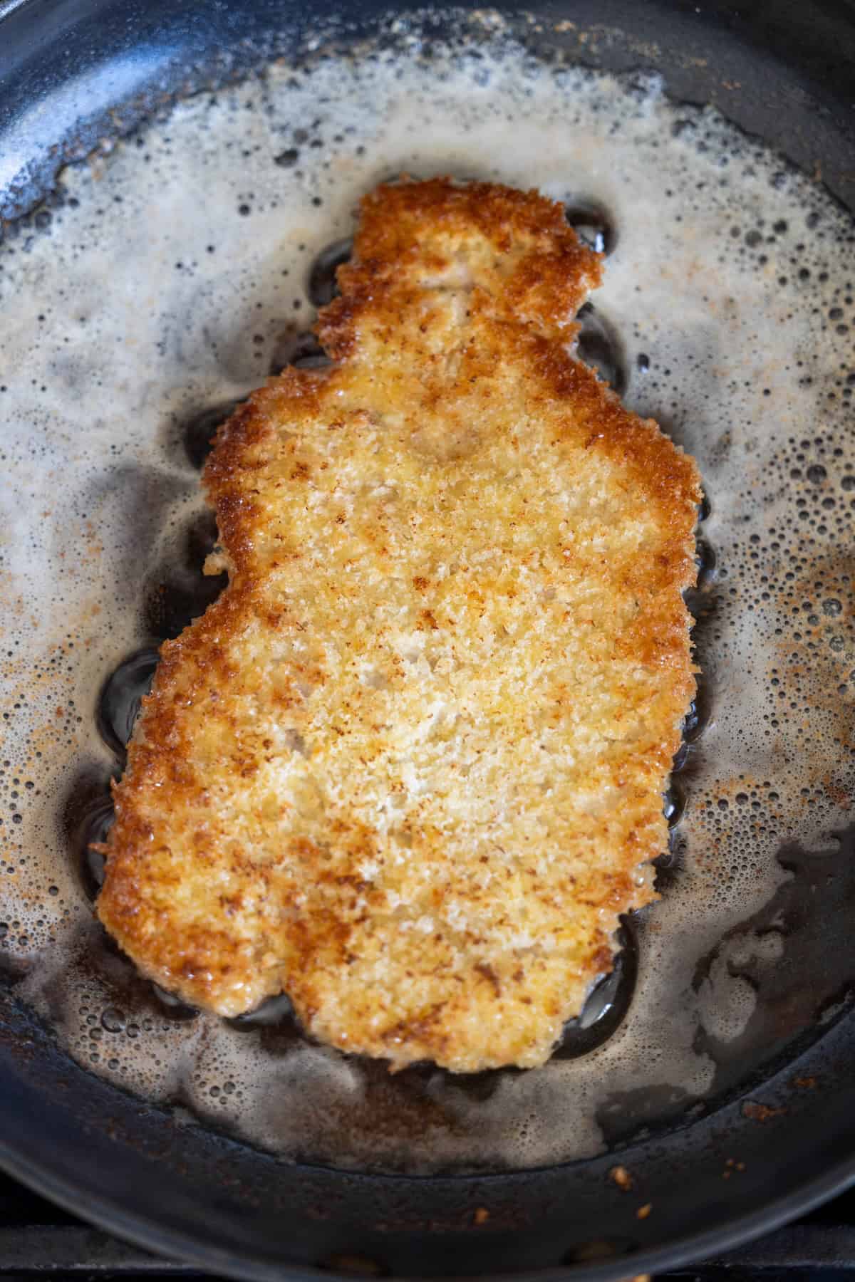 A breaded cutlet is being fried in a pan with bubbling oil and butter, revealing the golden brown crust typical of a classic Pork Schnitzel recipe.