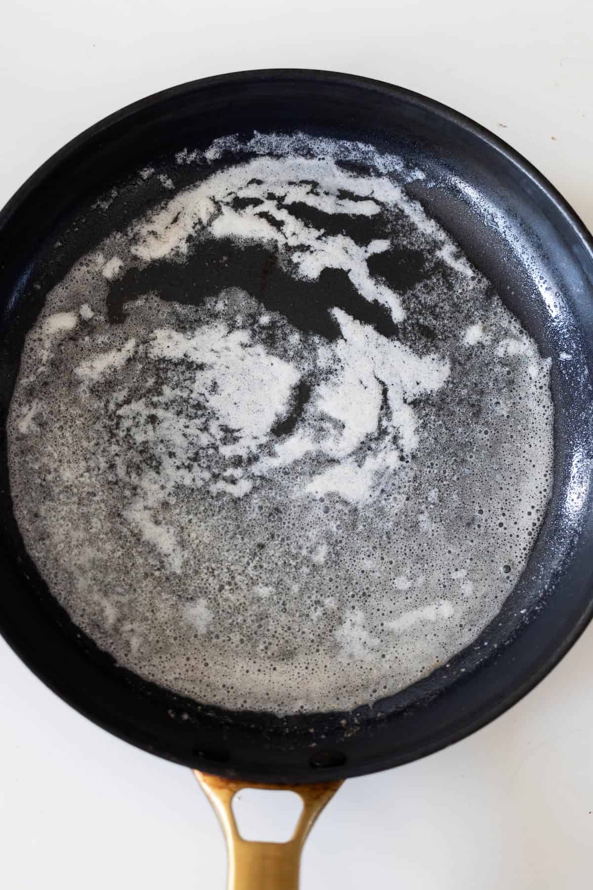 A black skillet with melted butter, forming white, frothy bubbles and streaks, sits on a white surface—perfect for starting a classic Pork Schnitzel recipe.