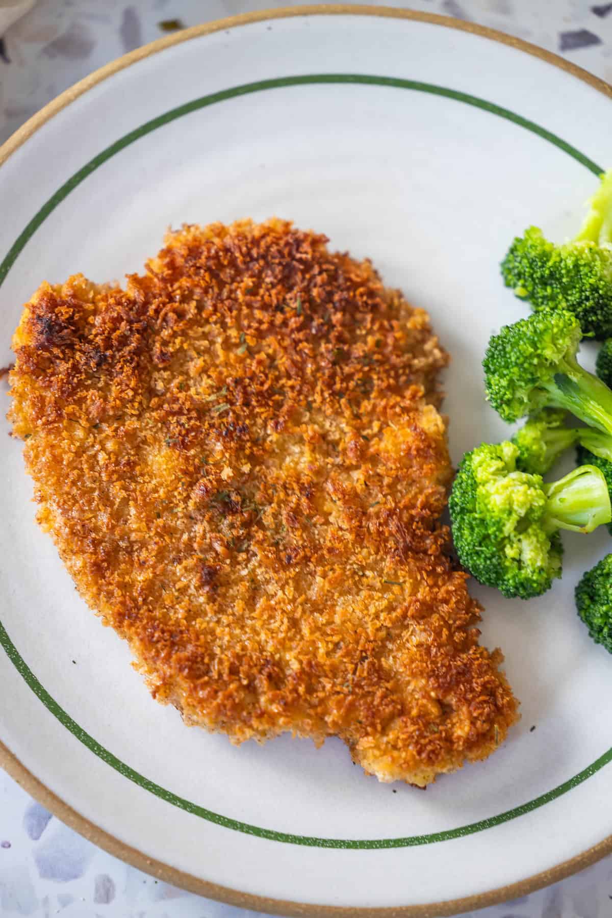 A crispy Pork Schnitzel is served on a plate alongside steamed broccoli florets—an easy and satisfying Pork Schnitzel recipe for any meal.