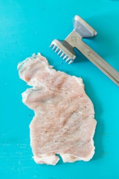 A raw chicken breast has been flattened with a meat tenderizer, ready for your favorite schnitzel recipe, both resting on a bright turquoise surface.
