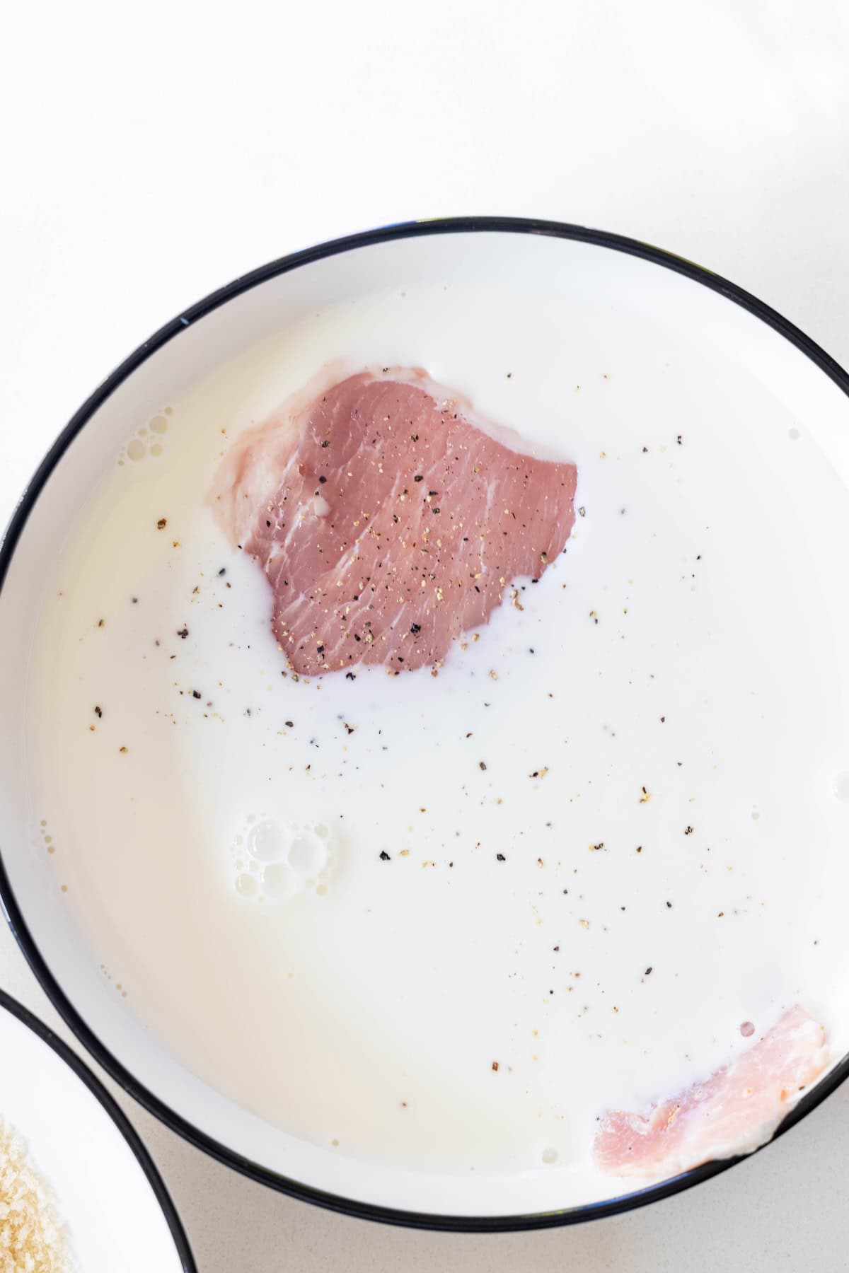 A raw pork chop soaking in a bowl of milk with black pepper sprinkled on top, perfect as the first step for a tender Pork Schnitzel recipe.