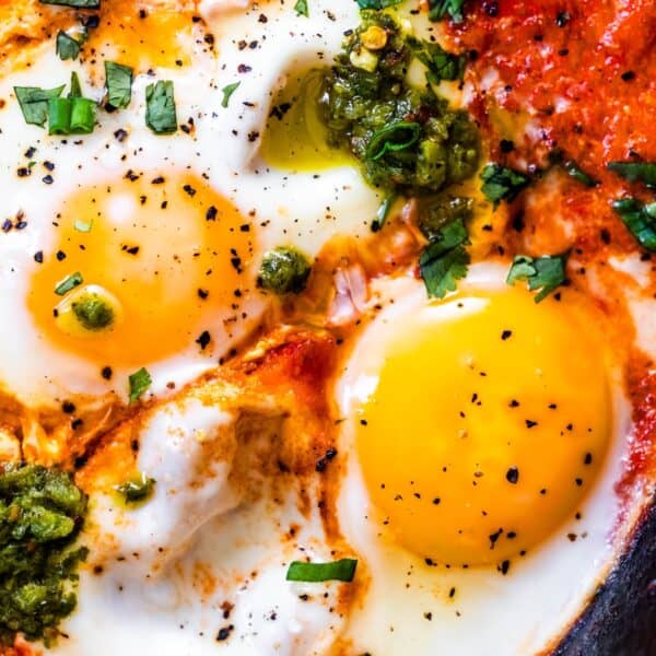 Close-up of three sunny-side-up eggs in a vibrant shakshuka, garnished with green herbs and pesto, seasoned with black pepper, and set in a rich red sauce—a classic Middle Eastern breakfast.