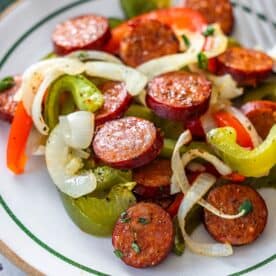 A plate of sliced sausage, sautéed onions, and bell peppers, seasoned with herbs and pepper, features this classic Sheetpan Kielbasa and Peppers dinner, served on a white dish with green trim.