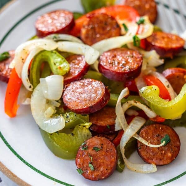 A plate of sliced sausage, sautéed onions, and bell peppers, seasoned with herbs and pepper, features this classic Sheetpan Kielbasa and Peppers dinner, served on a white dish with green trim.
