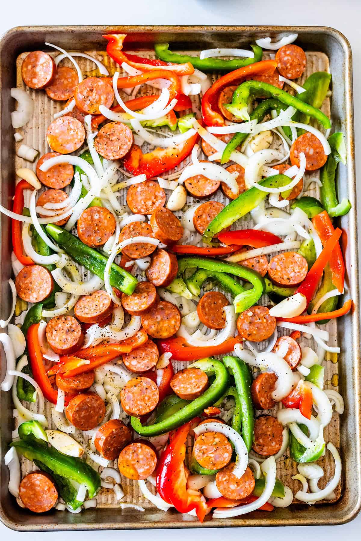 Sliced kielbasa, green and red bell peppers, and onions spread on a sheetpan lined with parchment paper, ready to be baked.