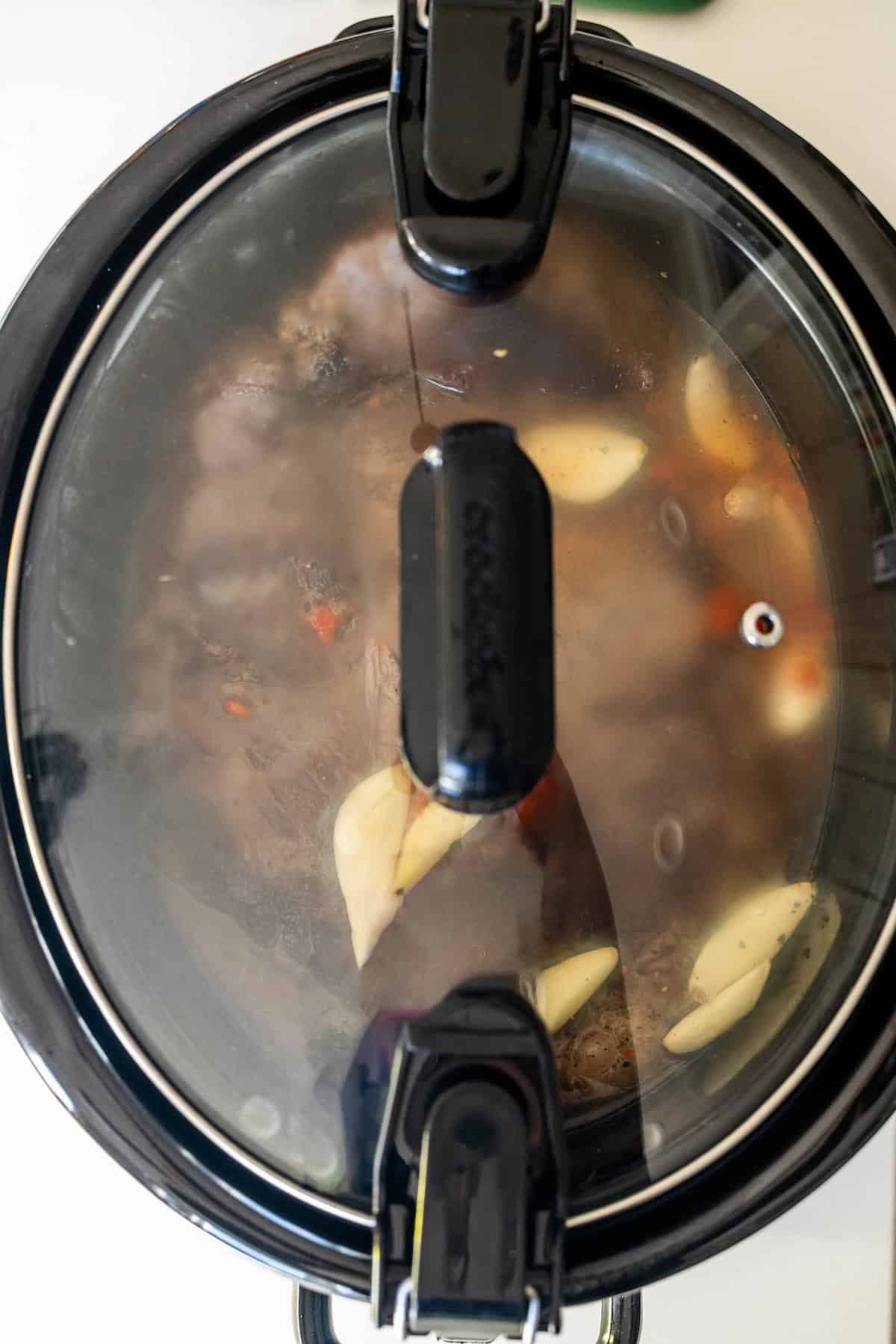 A slow cooker with a glass lid shows a chuck roast, garlic cloves, and broth inside, beginning the Slow Cooker Chuck Roast cooking process.