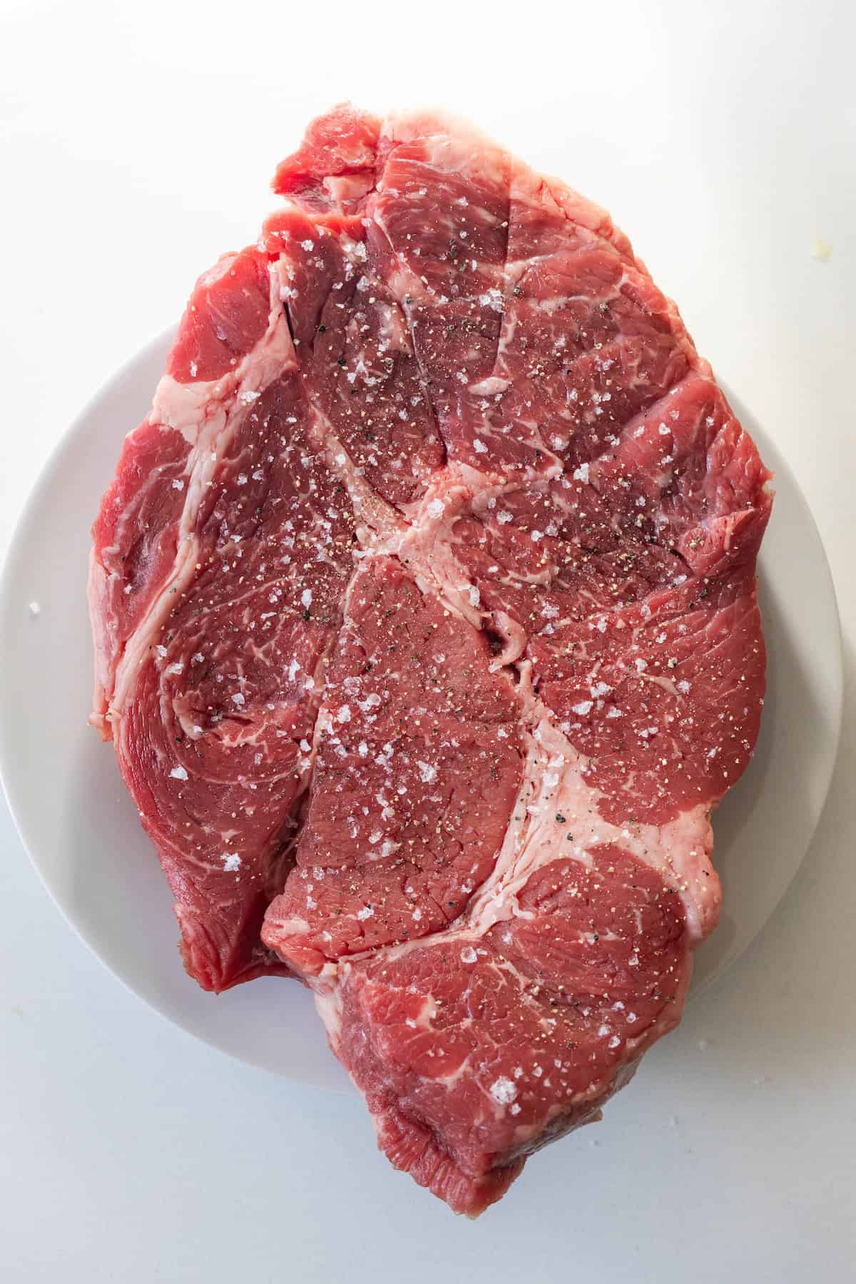 Raw beef steak on a white plate, sprinkled with coarse salt and cracked black pepper—perfect for preparing a Slow Cooker Chuck Roast, photographed on a white surface.