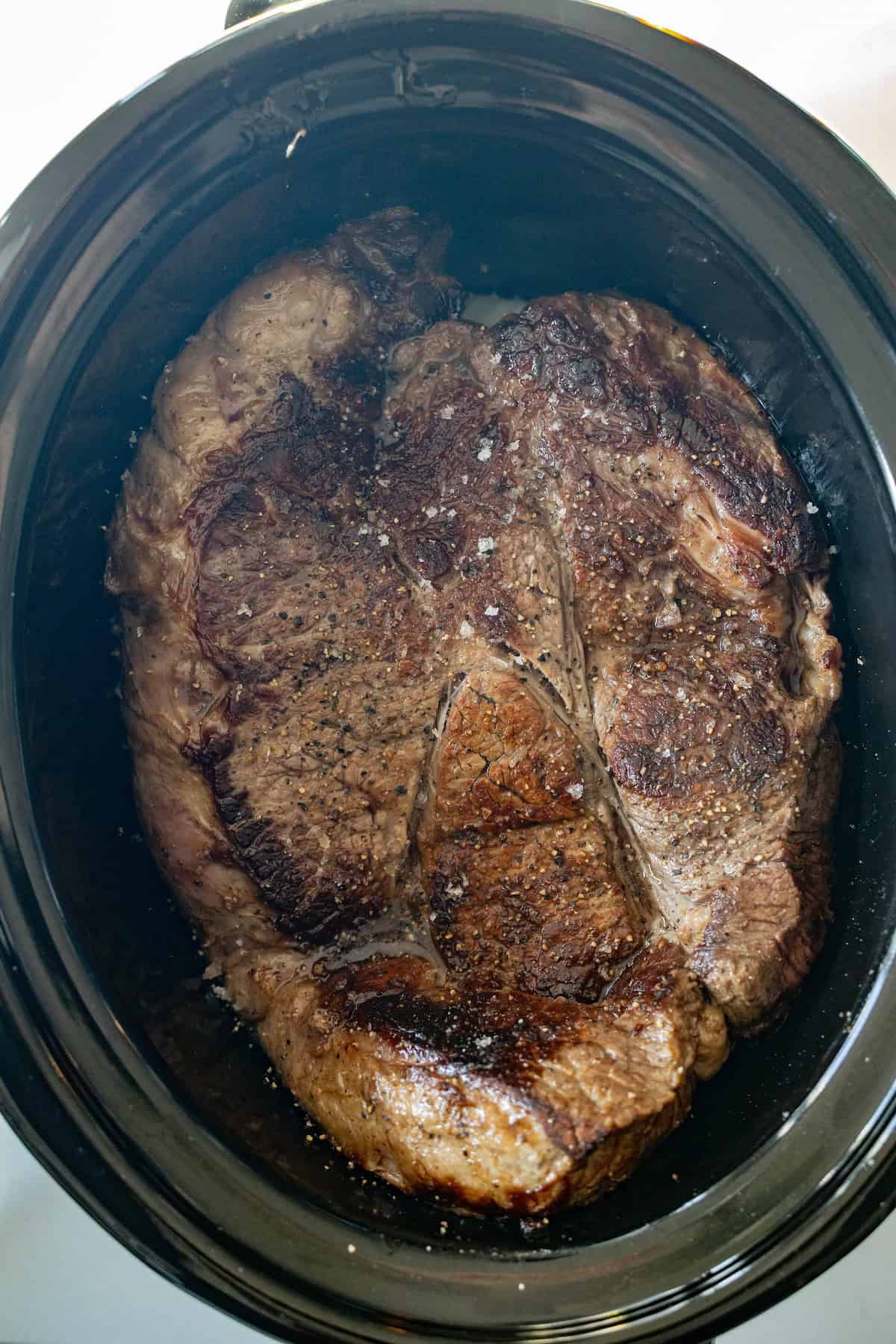 A large, browned slow cooker chuck roast sits in a black slow cooker, seasoned simply with salt and pepper.