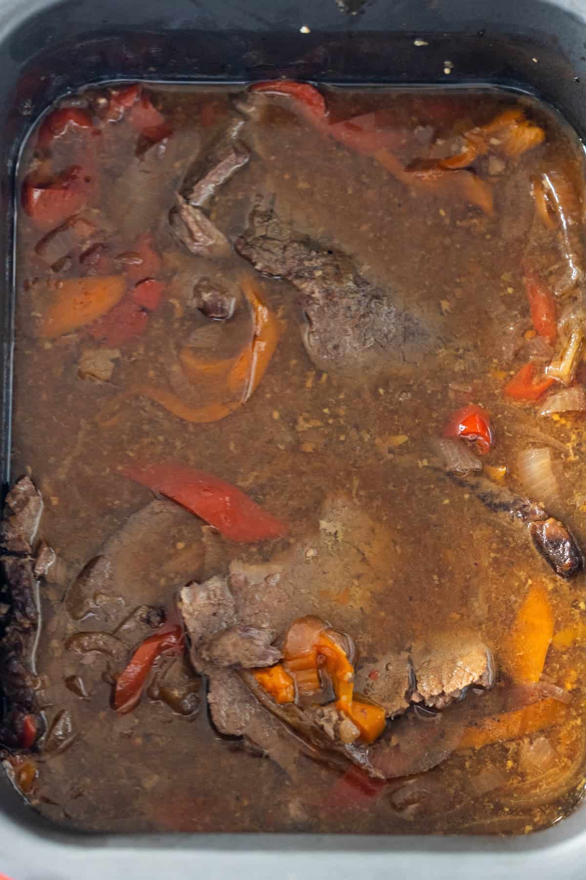 Slices of beef, bell peppers, and onions simmer in a brown broth inside a rectangular black slow cooker for an easy Philly Cheesesteaks-inspired meal.