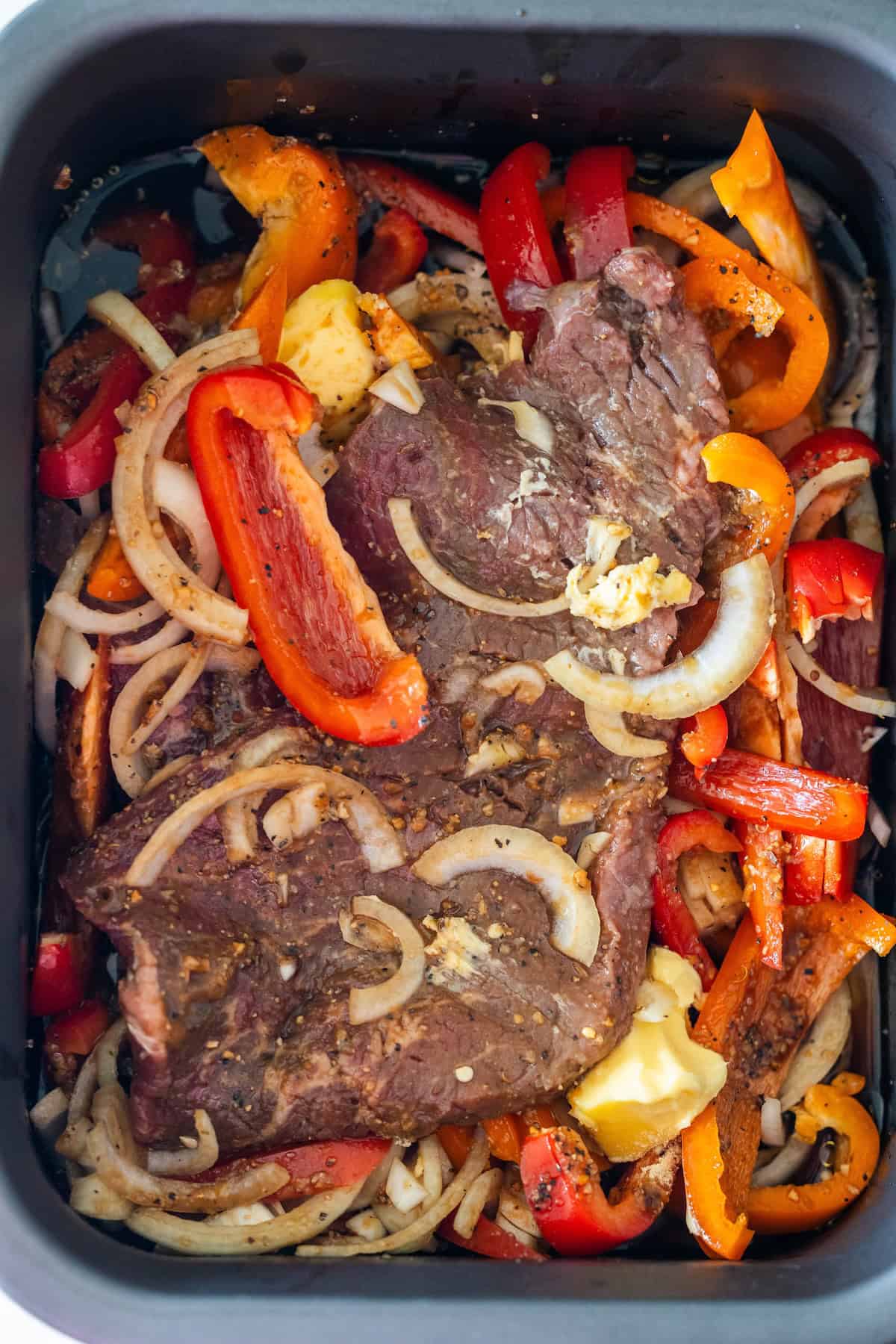Raw beef steak seasoned with spices, sliced onions, and red and orange bell peppers in a black cooking dish&mdash;perfect for a Slow Cooker Philly Cheesesteaks or your favorite cheesesteak recipe.