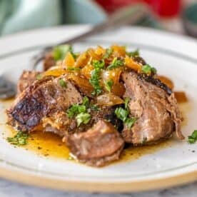 A plate of slow cooker steak garnished with chopped herbs and caramelized onions, served with sauce on a white plate.