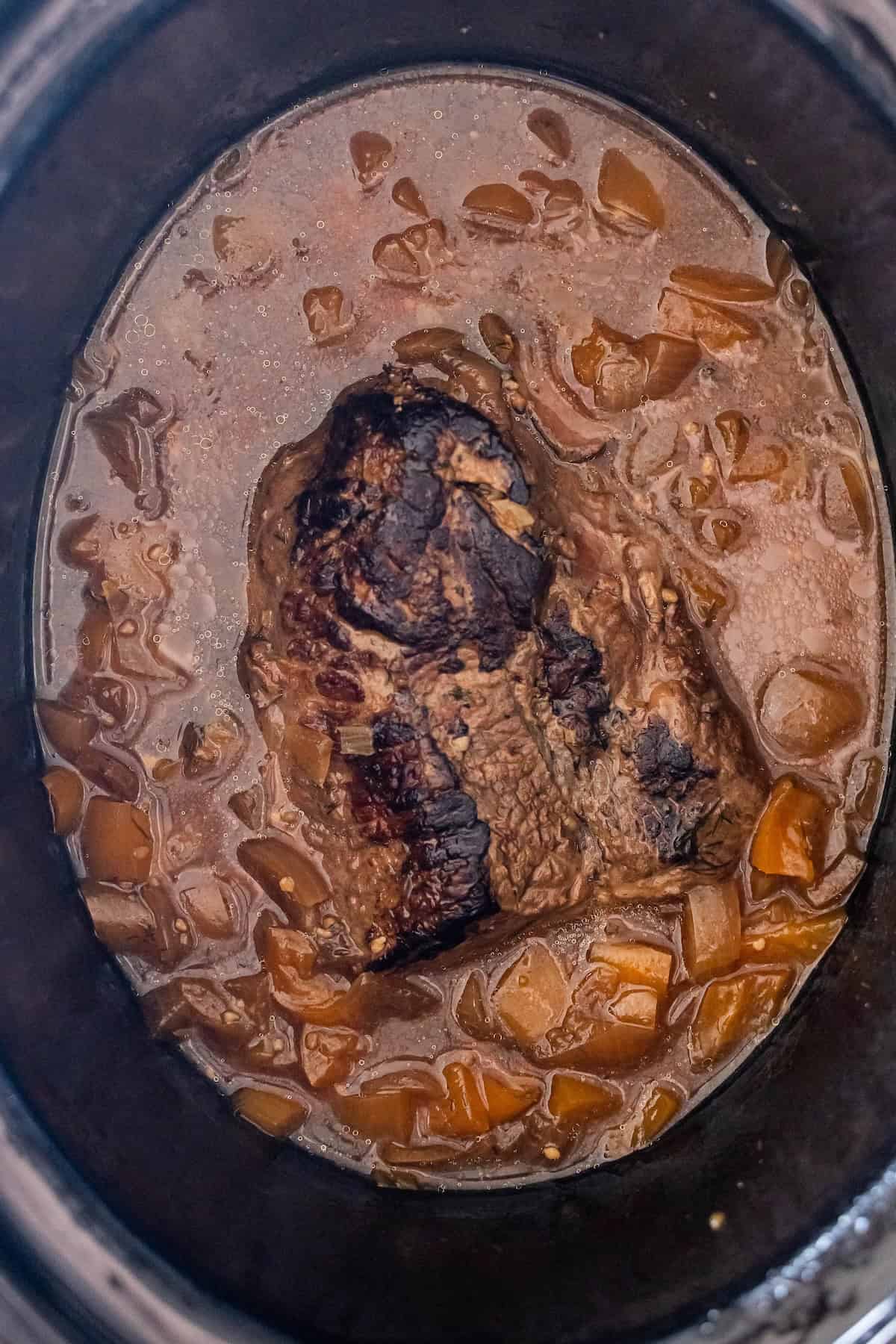 A cooked beef roast sits in a Slow Cooker with a brown sauce and chopped onions, offering tender flavor similar to Slow Cooker Steak.