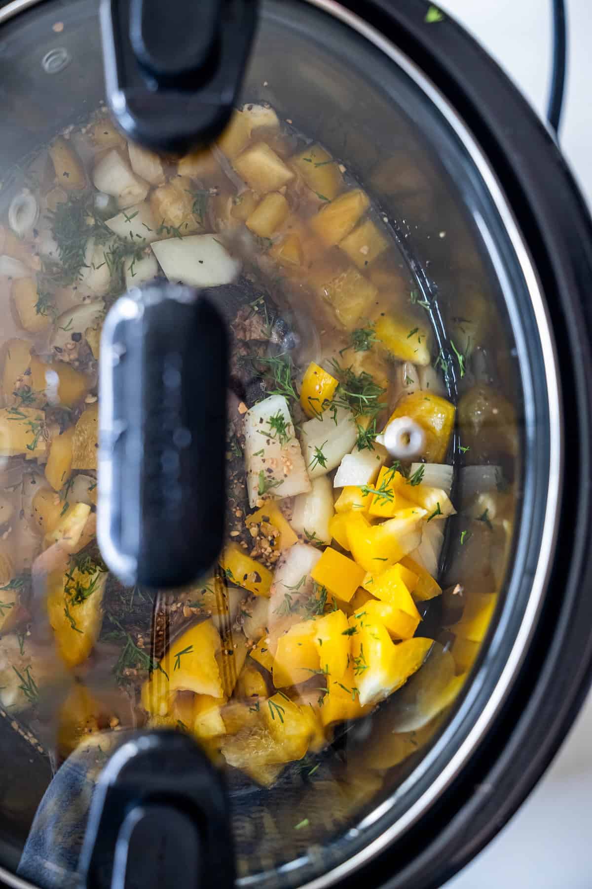 A slow cooker with a glass lid shows chopped yellow bell peppers, onions, herbs, and seasonings simmering inside—perfect for preparing tender Slow Cooker Sirloin Steak.