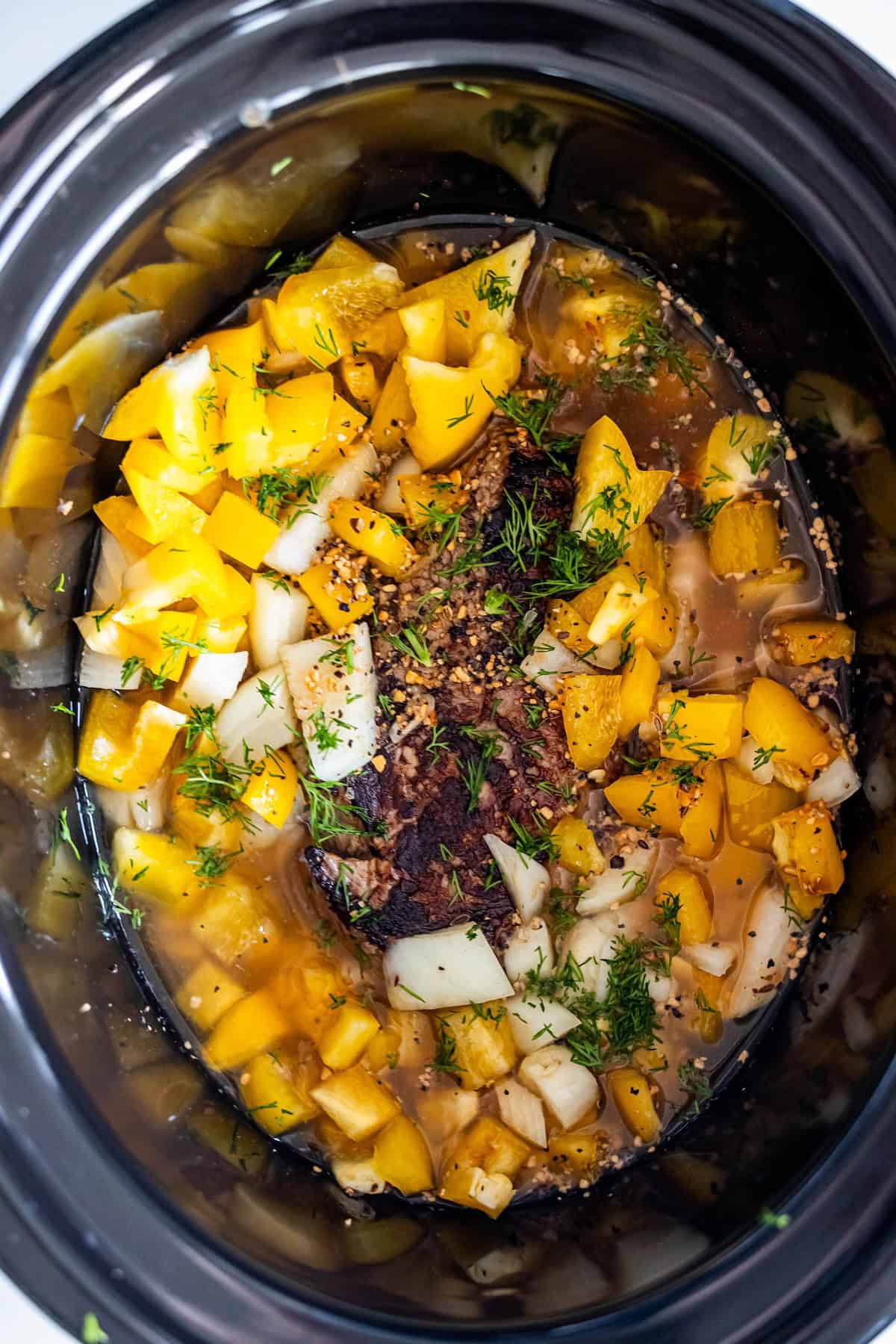 A slow cooker filled with seasoned sirloin steak, chopped yellow bell peppers, onions, herbs, and broth.