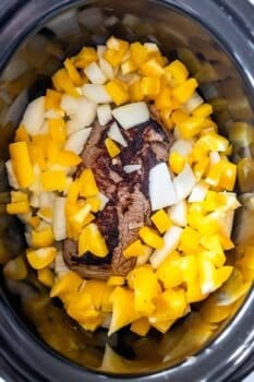 A piece of browned sirloin steak topped with chopped yellow bell peppers and onions inside a slow cooker.